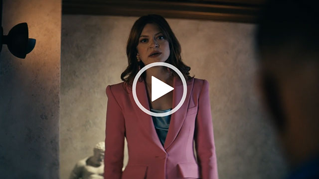 Colby Minifie stands in a stone wall room with black lamps and a dark wooden ceiling. She wears a structured pink blazer with a teal satin top.