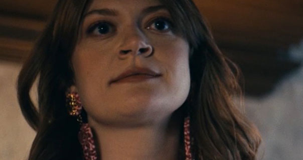 Colby Minifie is seen in a grey stone and dark wood room. She wears her long brown hair loose and layered. Cobie stars in a pair of large, dangling pink and purple jewel earrings.