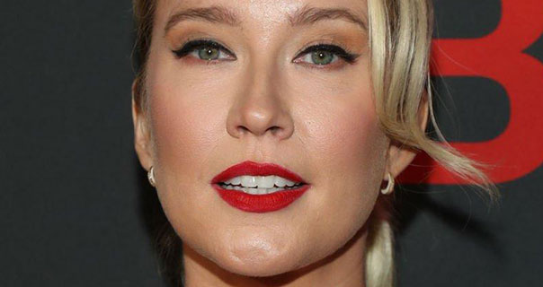 Anna Camp appears in a deep red matt lipstick and with her blonder hair tied back and up. She stands in front of a dark grey wall with red writing on it. Anna wears a pair of small gold hoop earrings and black winged eyeliner.