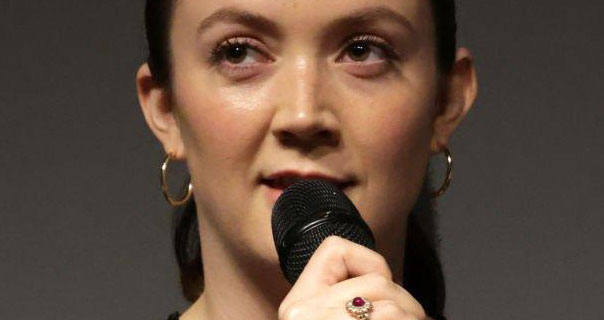 Billie Lourd appears in front of a dark grey wall. She wears her brown hair tied back. Billie holds up a microphone as she speaks on stage. She is seen in a pair of large thin gold hoop earrings and a pale pink lipstick.