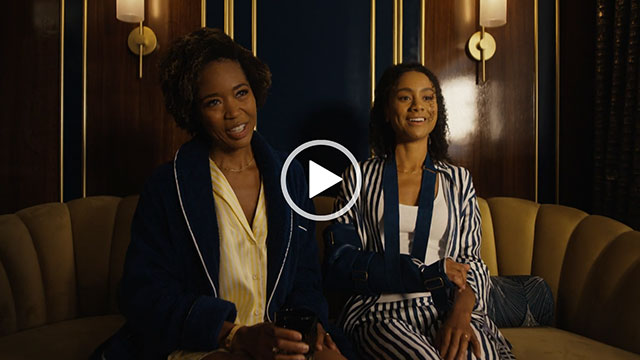 Marija Juliette Abney sits next to Michelle Núñez on a large dark olive colour couch. She wears a navy fluffy robe over yellow pyjamas. Michelle is seen in navy and white stripe sleepwear. They appear in ship cabin room that has dark wood with gold inlay and glowing lamps.