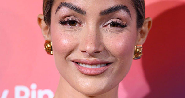 Frankie Bridge smiles as she looks into the camera. She wears a dark pink sheeny lipstick and heavy mascara and gold earrings. Frankie stands in front of a pale red wall with her shadow cast behind her. She has her thin brown hair tied up.