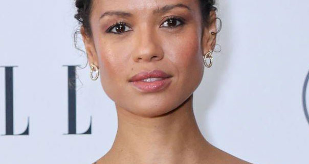 Gugu Mbatha-Raw is seen with her hair short hair tied up and in tight curls. She stands in front of a white wall that has black text on it. Gugu wears a pair of jewel studded hoop earrings. She appears in a gloss pink lipstick, black eyeliner and black mascara.