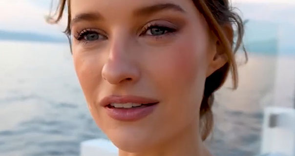 Victoria Magrath is seen wearing a dark pink lipstick with her hair tied back and up. She stands on the deck of a yacht with the sea and the setting sun behind her.