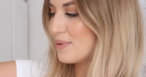 Katherine Bond is seen looking to the side. She holds one arm up as she stands in front of an off-white wall and wardrobe. Katherine wears a white t-shirt with her long blonde hair down and loose.