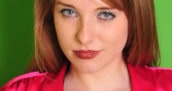 Jessica stands in front of a green screen in a studio. Her red hair is loose and pushed back behind her shoulders. She wears blue eye shadow, a dark red lipstick and a shiny pink top.