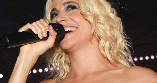 Pixie Lott sings on stage. She holds a black microphone close to her face as she performs. She wears her shoulder length blonde hair in tight waves as she stands on a dark stage with a row of small lights behind her.