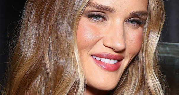 Rosie Huntington-Whiteley is seen smiling with her brown with blonde highlights down and loose. She wears a dark red gloss lipstick and pink blush against a dark background.