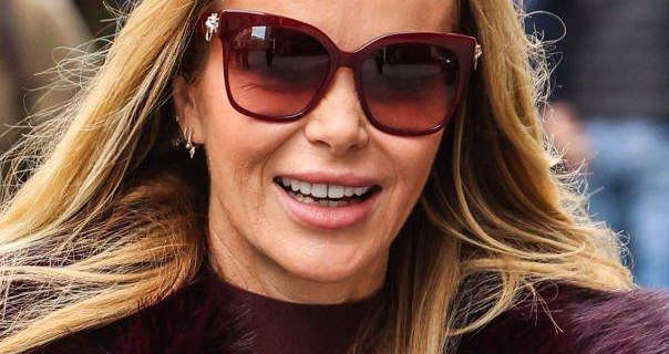 Amanda Holden is seen wearing a dark mauve top and fluffy mauve jacket. She has her blonde hair down. She appears in a pair of large sunglasses, silver earrings and a pink matte lipstick.