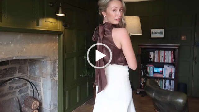 Josie Iron gathers her loose hair as she shows the bow design of the LilySilk top she has on. She stands next to a stone fireplace in front of a dark green wall and a wooden bookcase.