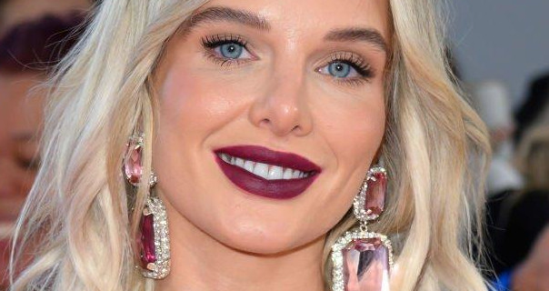 Helen Flanagan smiles as she appears on a red carpet with lots of fans behind her. She wears her blonde hair loose and a little messy. Helen is seen in a pair of large gemstone and diamond studded earrings and a dark red lipstick.