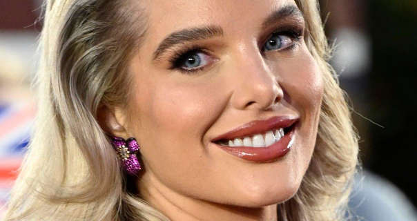 Helen Flanagan is seen smiling close-up. She has her thick blonde hair loose and around her shoulders. Helen wears black mascara with a dark red glossy lipstick and purple flower design earrings.