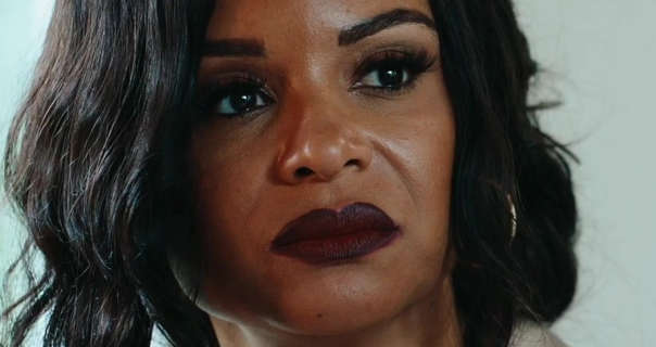 Joyful Drake is seen in front of a blue and white backdrop as she sits in a large living room. Her black hair frames her face. She wears a dark red lipstick and black mascara along with a pair of gold hoop earrings.