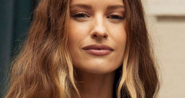 Victoria Magrath wears her hair in highlights, down and waved. She is seen in a dark pink lipstick as she stands on a street in Paris.