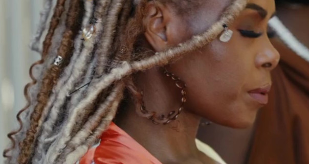 Chantal Maurice is seen from one side. She has her eyes closed. Chantal wears a pale red robe as she stands outdoors. Her brown and blonde hair is in dreadlocks.