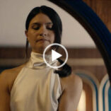 Phillipa Soo stands in a wooden arch inside a ship cabin. She wears a cream halter neck gown. Her black hair is gathered in a ponytail and she is seen in a dark pink lipstick and small stud earrings.