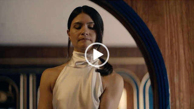 Phillipa Soo stands in a wooden arch inside a ship cabin. She wears a cream halter neck gown. Her black hair is gathered in a ponytail and she is seen in a dark pink lipstick and small stud earrings.