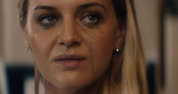 Kelsea Ballerini is seen with smudged mascara and makeup after crying. She wears her blonde hair down and pushed to one side. Kelsea appears in a beige and dark brown wood ships cabin room.