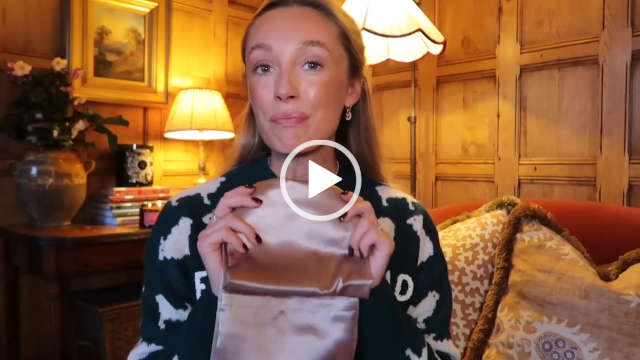 Josie Irons is seen wearing a green and white Christmas jumper. She sits in a corner of her wooden panel wall living room in yellow lamplight. Josie holds up a light bronze gold silk By Laima pillowcase.