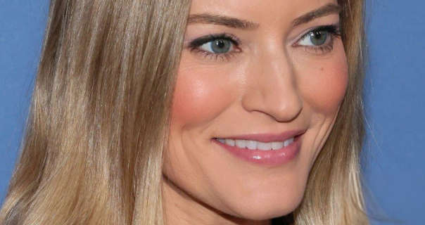 Justine Ezarik smiles and looks to one side as she poses for pictures. She wears her dark blonde hair straight and loose. Justine is seen in a pink blush, a glossy pink lipstick and black mascara as she stands in front of a dusty blue wall.