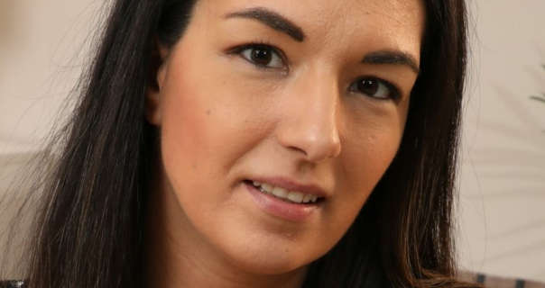 Rebecca G stars in front of a beige backdrop. She has her thick brown hair straightened and loose. Rebecca wears a dark pink matte lipstick with black eyeliner and black mascara.