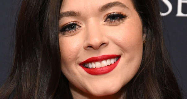 Sasha Pieterse wears a rich red matte lipstick as she stands in front of a dark grey wall with white writing on it. She has her black hair loose and down and slightly curled.