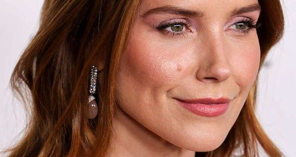 Sophia Bush is seen wearing light makeup and a dark pink lipstick. She stands in front of an off-white wall with her brown hair loose and in waves. Sophia is seen with dangling silver gemstone earrings.