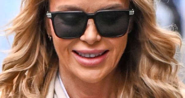 Amanda Holden smiles as she walks outside Global Studios in London. She wears her blonde hair curled and spread out. Amanda is seen in a dark pink lipstick and large black sunglasses.