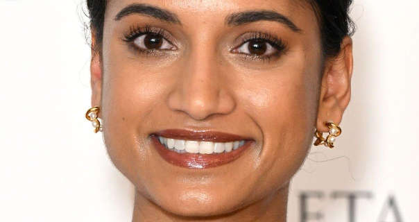 Amita Suman smiles while standing in front of a white wall with blurred black text on it. She wears her black hair tied up. Amita is seen in a dark red lipstick and a pair of gold pearl earrings.