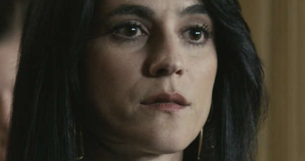 Carlye Tamaren stands in front of a light wood panel wall on a court room set. She has her black hair straight and loose. Carlye wears a dark pink lipstick and mascara with a pair of large gold chain earrings.