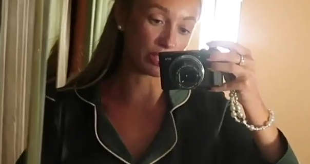 Josie Irons records herself in a full length mirror. She holds her black vlogging camera in one hand. Josie is seen with her long hair loose and pushed back. She wears pearl earrings and a pair of dark green silky pyjamas.