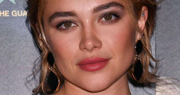 Florence Pugh is seen with her blonde cut short and styled in messy waves. She wears a pink lipstick and mascara with large earrings. Florence appears in front of a silver and black wall with gold text on it.