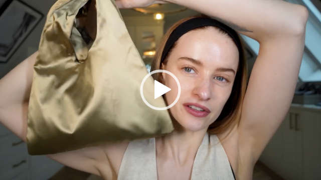 Victoria Magrath holds up a dark gold bag. She wears a black hair band to push back her loose hair. Victoria appears in her attic filming room at home with no makeup on.