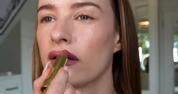 Victoria Magrath applies a dark red lipstick. She is seen in her white wood dressing room in front of a large mirror doors. Victoria has her hair down as she does her makeup.