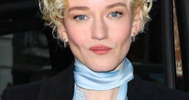 Julia Garner steps out of a large black SUV. She wears her blonde hair short and in tight curls. Julia is seen in a light blue top, black blazer and thin square earrings.