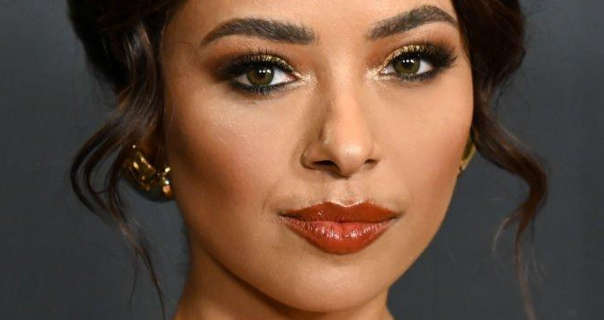 Kat Graham appears with her black hair tied up and with a few loose strands framing her face. She wears dark red lipstick, black eyeliner and mascara and a pair of large gold earrings. She appears in front of a dark grey wall.