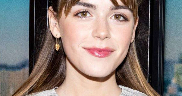 Kiernan Shipka stands in front of a black frame movie poster wall. She wears her light brown hair pushed back and with a long parted fringe. Kiernan is seen in a pink gloss lipstick, black mascara and a pair of dangling gold earrings.,