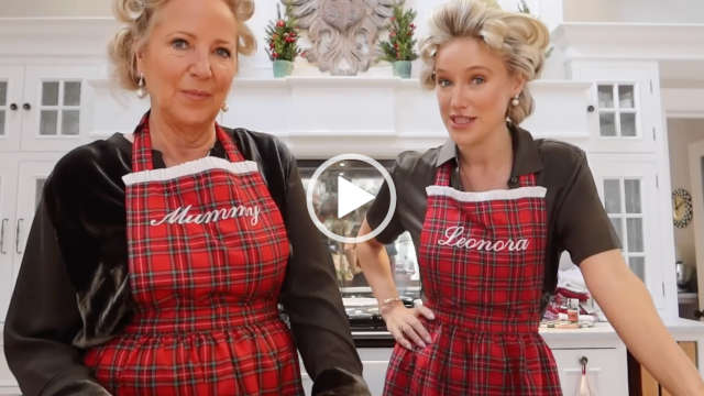 Leonora Smee stands next to her mother in the kitchen of her countryside home. Leonora wears her hair up in rollers and is seen in a dark green silk shirt dress, a tartan style apron and pearl jewellery.