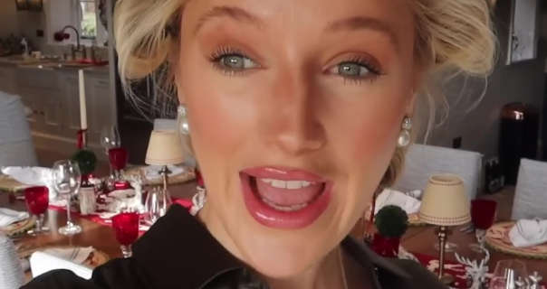 Leonora Smee stands in front of her large dinner table that is set for a festive gathering. She wears her blonde hair up in rollers. Leonora is seen in a forest green dress and a pink gloss lipstick.