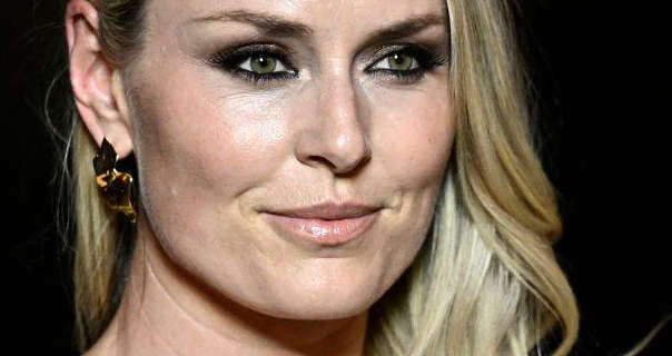 Lindsey Vonn is seen against a black background. She has her blonde hair wavy and down. Lindsey wears a dark pink lipstick with gold sheet earrings and black eye makeup.