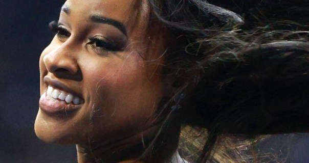 A Detriot Lions Cheerleader is seen with her black hair blowing backwards as she performs at a game. She wears a dark lipstick and thick lashes in front of a light grey backdrop.