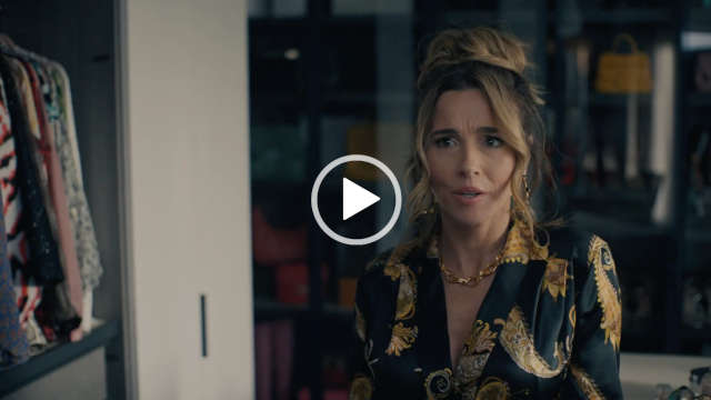 Linda Cardellini is seen wearing her hair up in a large bun. She appears in a black and gold print wrap nightwear top. Linda stands in a dressing room in front of large windows in partial darkness.