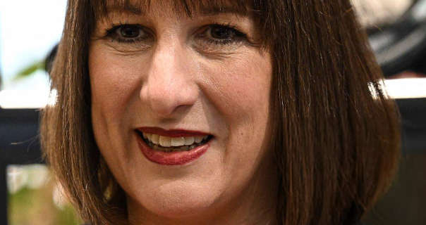 Rachel Reeves is seen smiling. She wears her short brown hair down and with a dark red lipstick. Rachel stands in front of a row of large black display shelves.