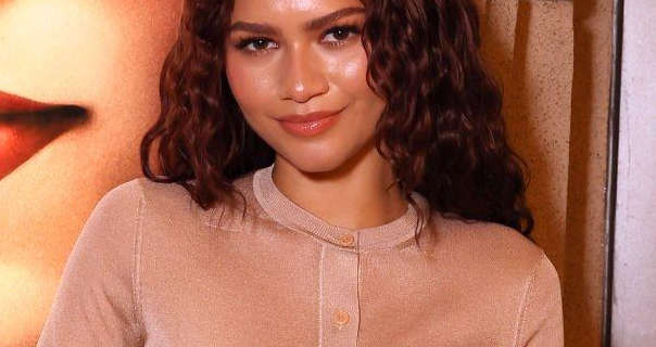 Zendaya stands in front of a poster for her film Challengers. She wears her hair curled and with light dewy makeup. Zendaya is seen in a dusty pink long sleeve cardigan.