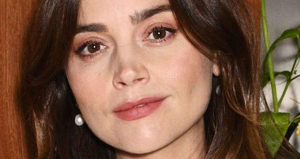 Jenna Coleman smiles a little as she looks into the camera. Her brown hair is loose and naturally wavy. She is seen wearing a pair of pearl drop earrings, mascara and a dark pink lipstick. Jenna stands in front of a wood panel wall and a green leafy plant.