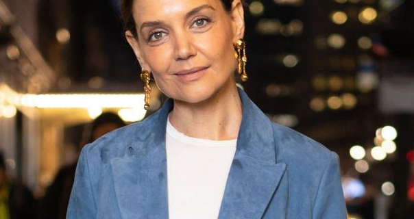 Katie Holmes wears a dusky blue blazer of a thin white top and hanging earrings. She is seen with her black hair tied up. Katie stands on a New York City street at night with a sky scraper and bright lights behind her.