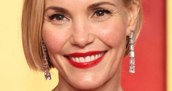 Leslie Bibb stands in front of a pale yellow and pastel red wall. She wears her blonde hair cut short. Leslie is seen in a dark red gloss lipstick and large hanging square gemstone earrings.