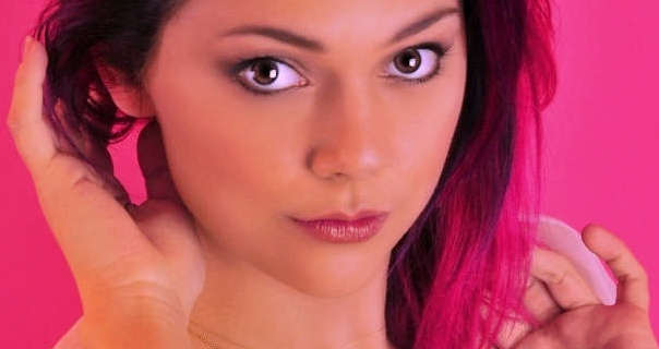 Polly is seen against a pink studio screen background. Her black pink tints hair is pushed to one side. Polly wears black mascara and a dark pink gloss lipstick. She touches her hair with the fingers of both her hands as she looks directly into the camera.