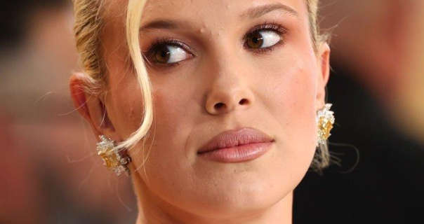 Millie Bobby Brown looks to one side during a red carpet event. She wears her hair coloured blonde and tied up with a strand pulled in front of her face. Millie is seen in a dark pink lipstick and a pair of large gold and silver earrings.