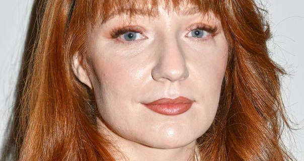 Nicola Roberts is seen in sharp focus in front of an off white wall. She wears her red hair down, loose and a little messy. Nicola is seen in a matt red lipstick, orange eye shadow and volumised eyelashes.